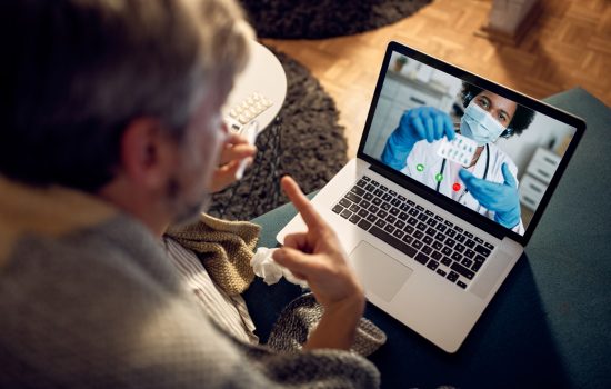 Close-up of man with fever consulting his doctor during a video call at night from home. Focus is on female doctor on laptop screen.